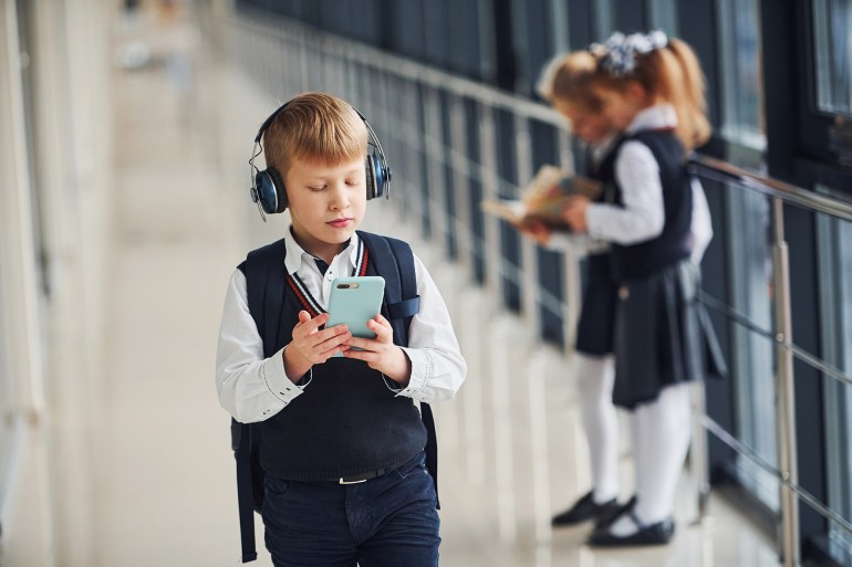 Little boy with phone and headphones standing in front of school kids in uniform that together in corridor. Conception of education.