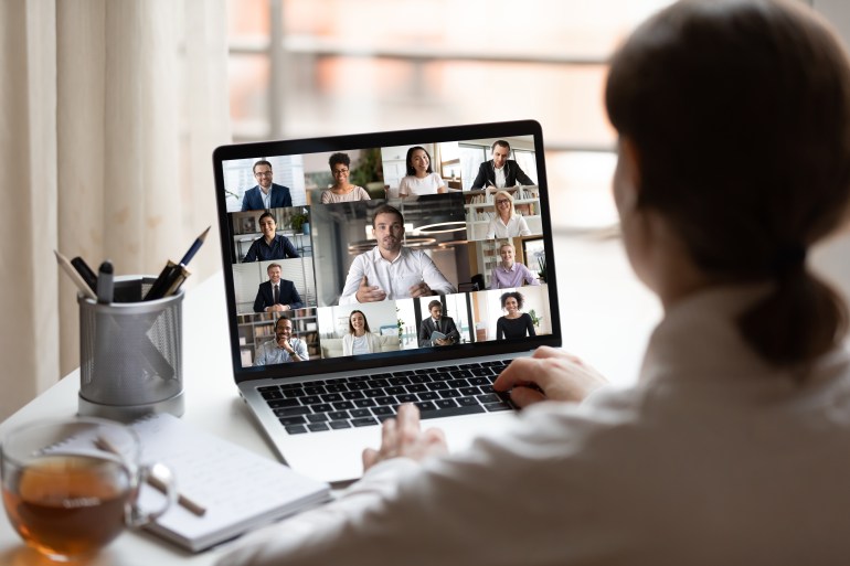 View over businesslady shoulder seated at workplace desk look at computer screen where collage of many diverse people involved at video conference negotiations activity, modern app tech usage concept