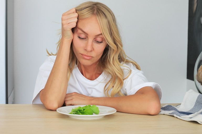 I'm fed up with untasty disgusting salad. Close up unhappy unsatisfied grimacing sad upset lady looking down at plate of lettuce on table.