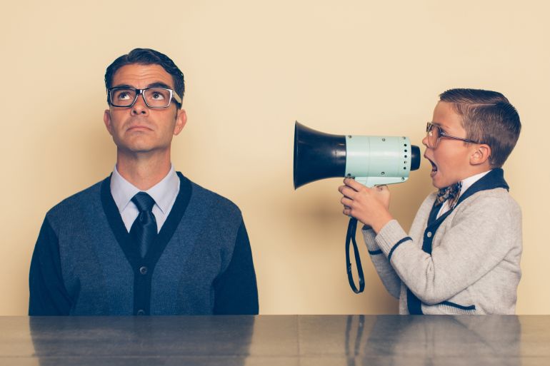 A young nerd boy in eyeglasses and a bow tie is yelling at his dad through a megaphone. His dad is looking up at the ceiling and ignoring what the son is saying. The son is frustrated with his dad because he won't listen as there is a generation gap غيتي